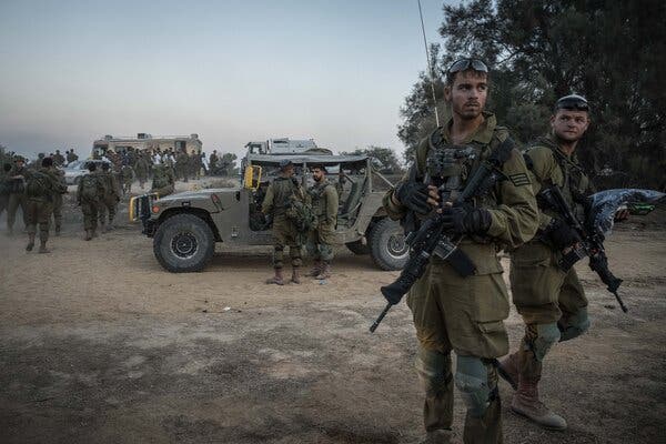 Armed Israeli soldiers stand on muddy ground and near military vehicles.