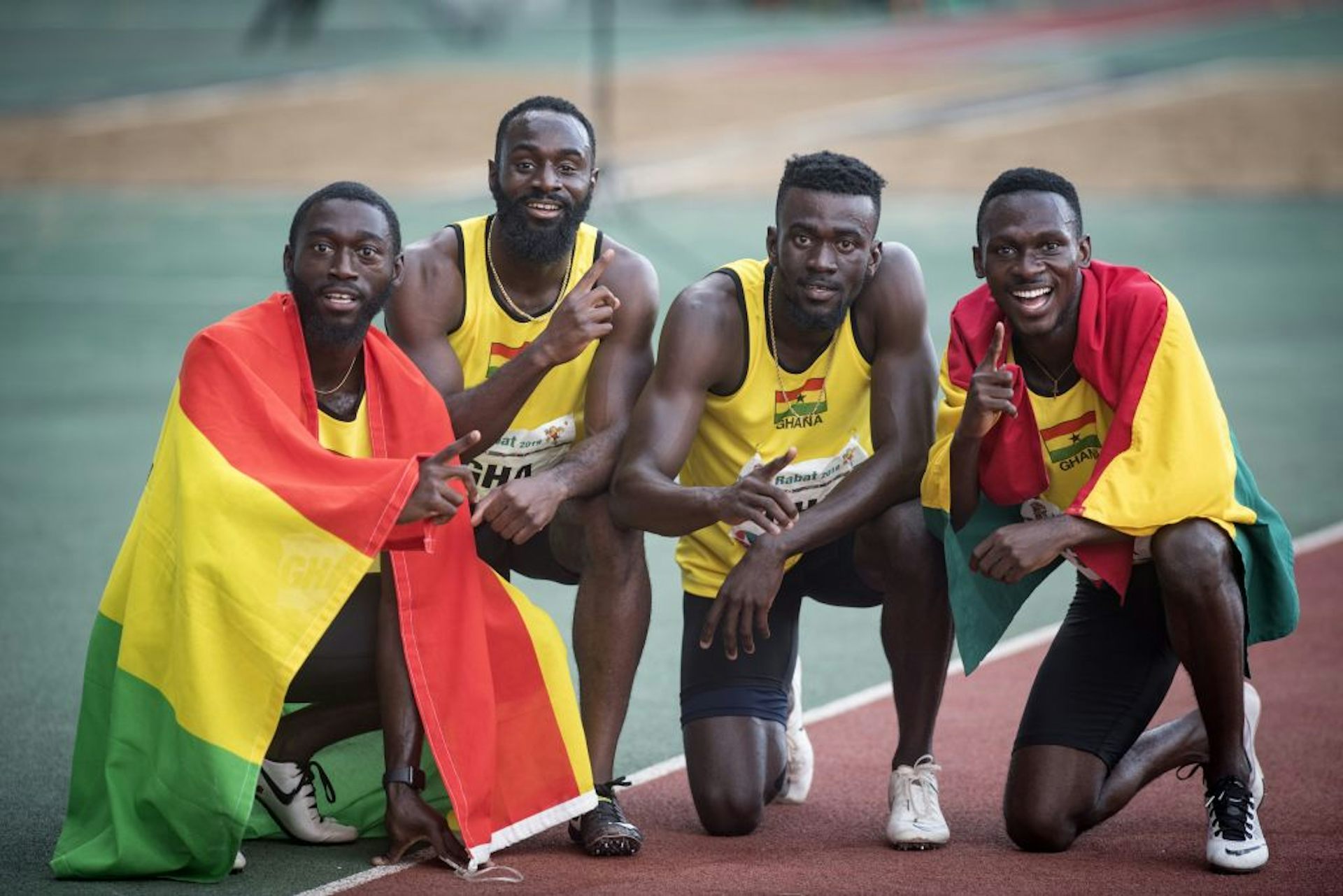 Four men in athletics gear kneel and crouch on a running track, two of them draped in the green, red and yellow flags of Ghana.