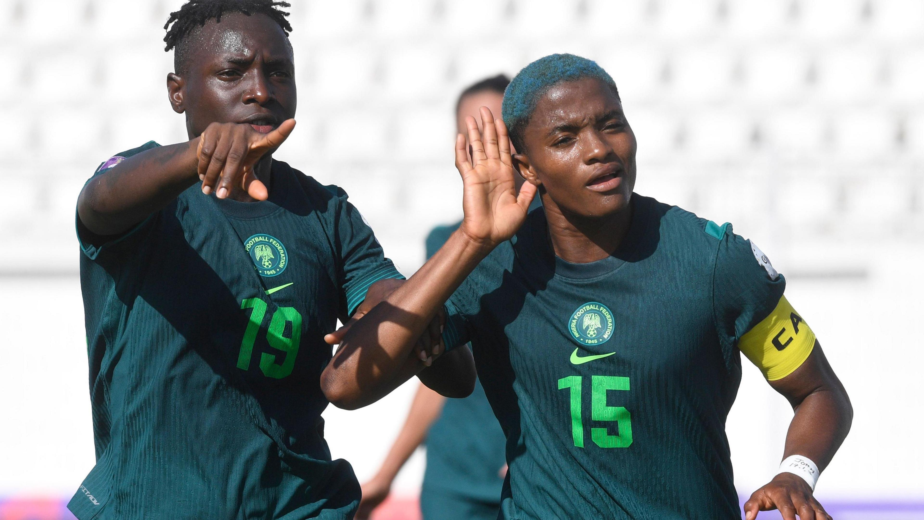 Rasheedat Ajibade in dark green Nigeria kit and with dyed blue hair celebrates by cupping her right hand to her ear while team-mate Chinwendu Ihezuo grabs that same arm and points with her free hand