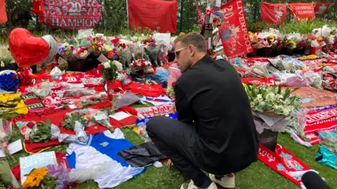 Marc Gaier/BBC Jordan Henderson, in a black t-shirt and navy blue trousers, sits on his haunches in front of a sea of tributes including football kits, flowers, flags, scarves and balloons.