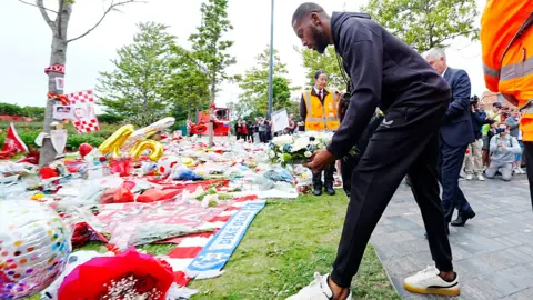 PA Media Beto, in a navy blue tracksuit, places a wreath made up of white flowers on to a grassy area containing colourful tributes including bouquets, scarves, balloons and flags.