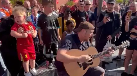 Phil Munns/BBC Mark Phillip sits on a stone bench playing an acoustic guitar surrounded by a crowd of people, some recording on their phones. A little blonde-haired boy in a full replica Liverpool kit is being held by a woman behind him.