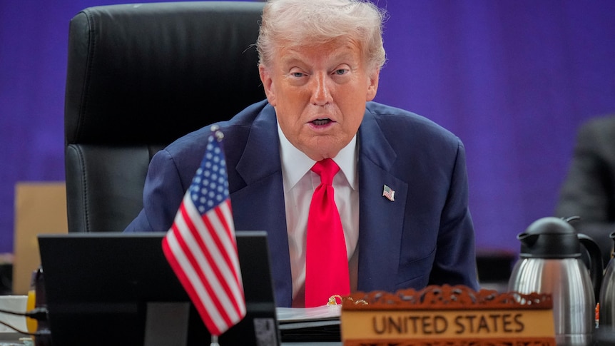 Donald Trump, wearing a suit and red tie, speaks behind a sign that says United States.